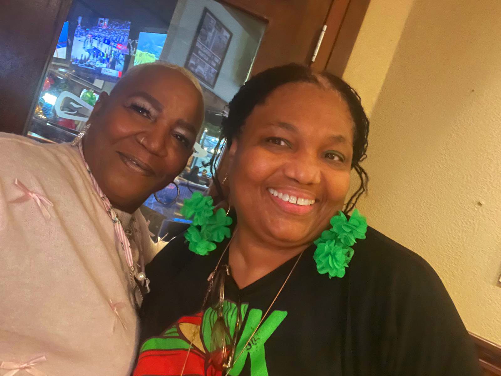 Two women smiling indoors, one wearing green earrings and a black shirt with red and green design.